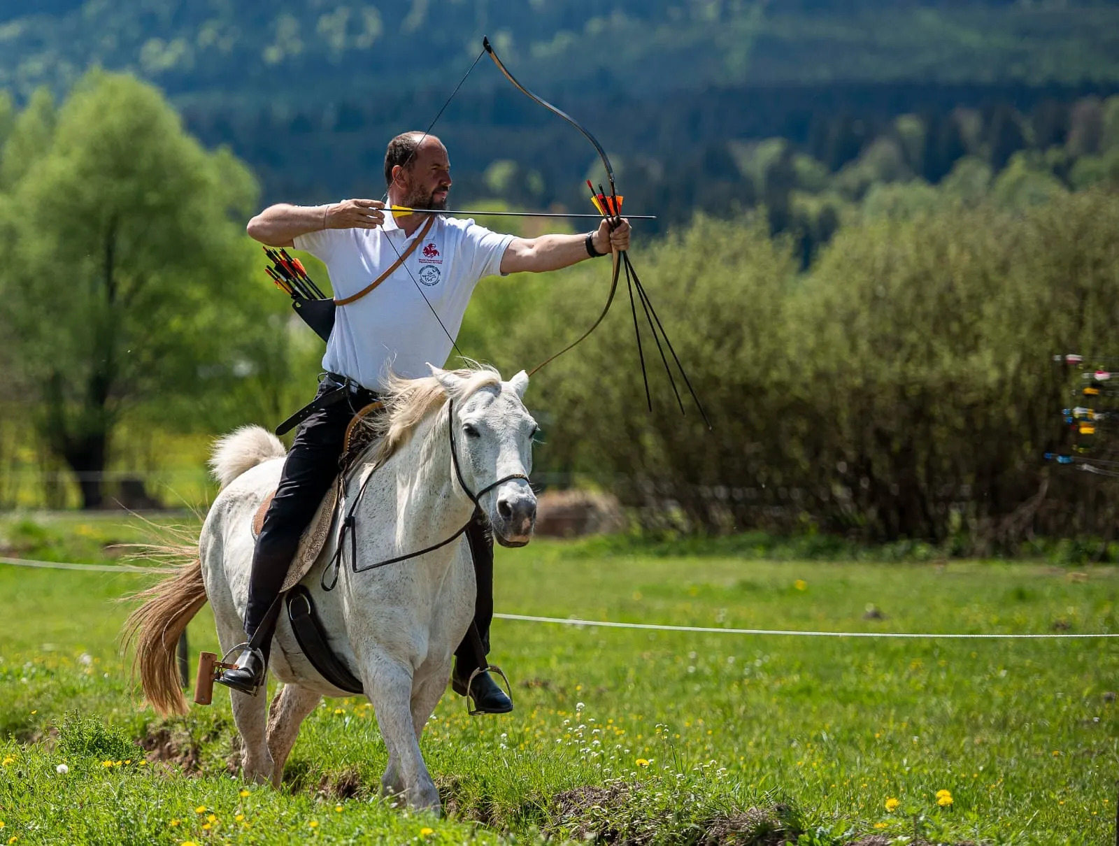 Un arcaș cu arcul întins pe un cal alb, într-o pajiște sub munți.